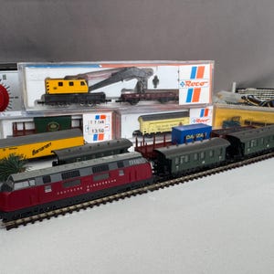 Puede incluir: Un set de tren a escala con una locomotora Deutsche Bundesbahn de color granate tirando de varios vagones de pasajeros verdes, junto a vagones de carga como uno amarillo Bananien y otro azul, además de cajas de embalaje de la marca Roco.