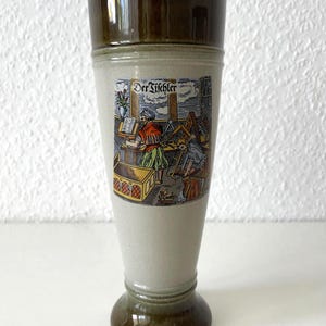 Pode incluir: Um copo de cerveja alto de cerâmica com a parte superior e a base marrons e um corpo cinza claro. O corpo apresenta uma ilustração colorida de uma oficina de carpinteiro com o texto "Der Tischler".