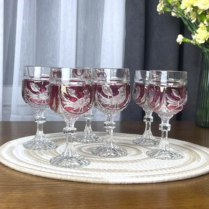 Puede incluir: Un juego de seis copas de vino de cristal transparente con una superposición de color rojo arándano intenso. Cada copa tiene un tallo facetado y una base con un diseño grabado detallado de un pájaro posado entre hojas.