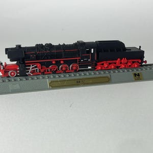 Peut inclure: Une locomotive à vapeur miniature noire et rouge, identifiée comme BR-52, montée sur un socle de présentation en plastique gris représentant une voie ferrée. Le modèle présente des roues détaillées, un tender et une plaque dorée avec le texte BR-52.