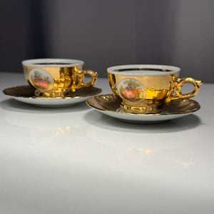 May include: Two gold-colored teacups and saucers. The cups and saucers have a gold trim and a small painted scene on the cup. The handles are ornate. The saucers are round and gold-colored.