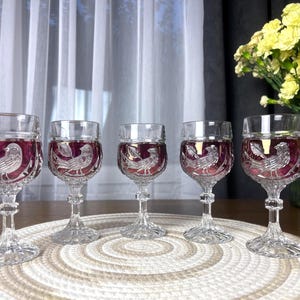 Può includere: Un set di cinque calici in vetro trasparente con una coppa color magenta intenso e un design a uccello inciso. Ogni bicchiere presenta uno stelo sfaccettato e una base strutturata, che mostra un rilievo dettagliato di un uccello.