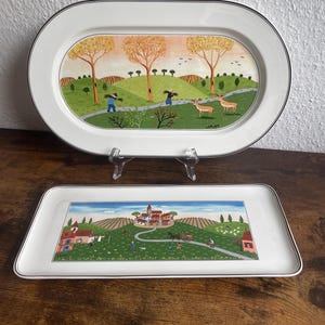 Può includere: Un piatto da portata ovale e un piatto rettangolare in ceramica, entrambi con bordi bianchi e un sottile profilo nero. Il piatto ovale raffigura un paesaggio con alberi, due cervi e figure. Il piatto rettangolare mostra una scena di villaggio.