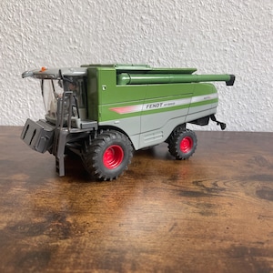 May include: A green and grey Fendt Hybrid combine harvester toy model featuring a detailed driver cabin, large black tires with red rims, and a long side-mounted unloading auger. The side of the vehicle displays the Fendt Hybrid brand name in white.