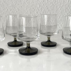 May include: A set of six clear glass goblets with a modern design. Each goblet has a short, dark gray stem and a wide, slightly tapered bowl. The glasses are arranged on a white surface against a white background.