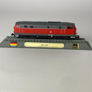 Könnte beinhalten: Eine rote und graue Modell-Diesellokomotive mit der Aufschrift BR-218, montiert auf einem grauen Gleissockel mit deutscher Flagge, einem goldenen Namensschild und einem quadratischen N-Skala 1:160 Spur 9mm Etikett auf der rechten Seite.