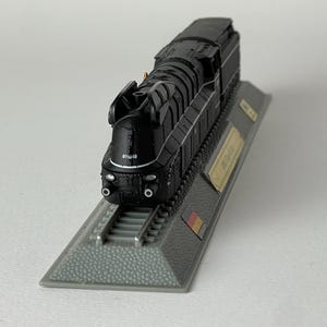 May include: A black model steam locomotive featuring the number 011018 printed on the front. The train is mounted on a gray textured display base that includes a small German flag emblem and a gold-colored identification plaque on the side.