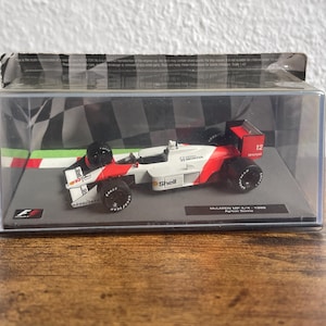 Könnte beinhalten: Ein 1:43 Modell des McLaren MP 4/4 Formel-1-Rennwagens von 1988, gefahren von Ayrton Senna. Das weiß-rote Auto trägt Shell- und Honda-Logos, schwarze Reifen und die Nummer 12 und ist in einer durchsichtigen Kunststoffvitrine mit Zielflaggenmuster.