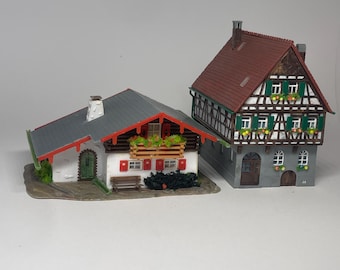 Op de afbeelding: Twee miniatuur modelgebouwen. De linker structuur is een huis met één verdieping, een grijs dak, witte muren, rode luiken en een balkon met groene planten. De rechter structuur is een hoger, meerverdiepingsgebouw met een bruin dak en vakwerk.