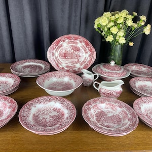 Op de afbeelding: Een servies van rood en wit transferware met gedetailleerde landelijke scènes. De set bevat een grote ovale schaal, diverse kommen, borden, een terrine met deksel en twee roomkannetjes, allemaal versierd met ingewikkelde bloemmotieven.