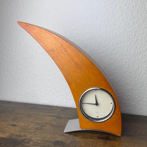 Op de afbeelding: Een gebogen, halvemaanvormige bureauklok van hout met een warme, lichtbruine houtnerfafwerking. De klok heeft een ronde, gebroken witte wijzerplaat met zwarte wijzers en een zilverkleurige metalen rand, geplaatst op een zilverkleurige metalen voet.