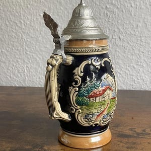 Może przedstawiać: Ceramiczny kufel do piwa z ciemnoniebieskim korpusem ozdobionym kolorowym reliefem przedstawiającym górski krajobraz z domem i drzewami. Posiada jasnobrązową podstawę i obrzeże, ozdobne kremowe ucho oraz spiczastą, metalową pokrywkę z tłoczeniami.