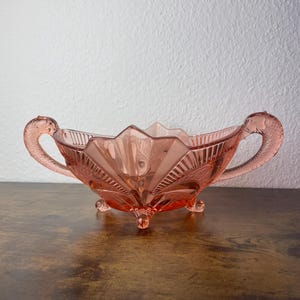 Puede incluir: Un cuenco de vidrio prensado rosa con un borde festoneado y dos asas con forma de delfín. El cuenco se apoya sobre tres pies pequeños y presenta un patrón texturizado en forma de abanico que irradia desde el centro hacia los bordes.