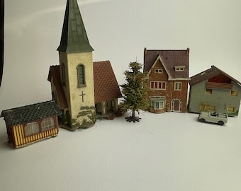 Op de afbeelding: Een verzameling miniatuurmodelgebouwen waaronder een geel schuurtje, een kerk met een hoge toren en kruis, een bruin huis, een groen huis, een kleine groenblijvende boom en een piepklein grijs voertuig op een effen witte ondergrond.