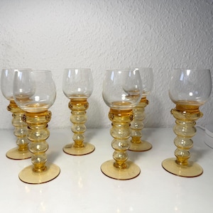 May include: A set of six vintage wine glasses. Each glass features a clear bowl atop a stem with a series of amber-colored, rounded segments. The base and the decorative ring at the top of the stem are also amber-colored. The glasses are arranged on a white surface.
