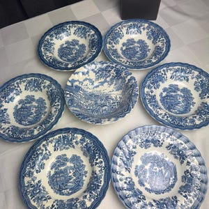 Può includere: Un set di sei ciotole da zuppa rotonde in ceramica con un motivo blu e bianco. Ogni ciotola mostra una scena paesaggistica dettagliata con edifici e alberi, circondata da un bordo floreale su un bordo smerlato.
