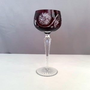 Può includere: Un calice da vino in cristallo tagliato rosso intenso con un intricato motivo a stella e foglie inciso sulla coppa. Lo stelo è sfaccettato e trasparente, e scende verso una base circolare con un disegno a stella radiante inciso.