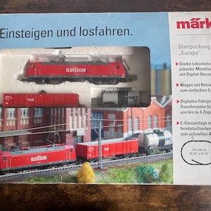 Pode incluir: Uma caixa de conjunto inicial de comboio em miniatura Märklin H0, modelo Europa 29130. A embalagem apresenta uma locomotiva elétrica vermelha Railion, um vagão de carga vermelho e um vagão-tanque. A caixa exibe o texto Einsteigen und losfahren e um diagrama de via de 115 cm por 85 cm.