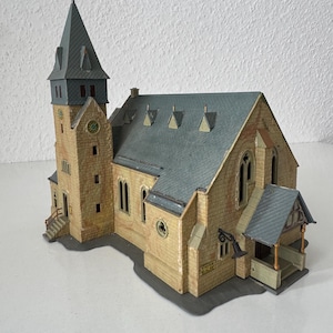 Könnte beinhalten: Ein Kirchenmodell mit beigefarbenen, steinstrukturierten Wänden, einem dunkelgrauen Satteldach mit mehreren Dachgauben und einem hohen Kirchturm. Das Gebäude verfügt über Rundbogenfenster, einen kleinen Seiteneingang und ein Ziffernblatt.
