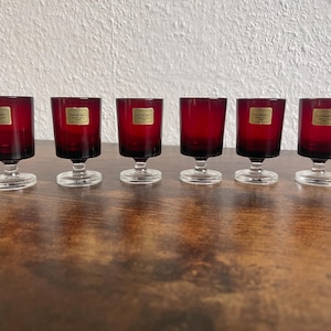 Può includere: Un set di sei piccoli calici in vetro rosso scuro con stelo e base in vetro trasparente. Ogni calice presenta un adesivo rettangolare color oro sulla parte anteriore con il testo Luminarc France stampato in nero.