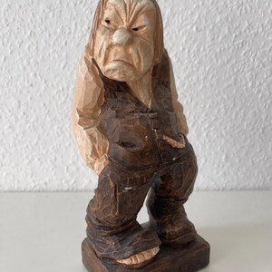 Könnte beinhalten: Eine handgeschnitzte Holzfigur eines mürrisch aussehenden Charakters mit hellem Gesicht und dunkelbrauner Kleidung. Die Figur steht auf einem rechteckigen Sockel, in dessen Vorderseite "Sier Karten" eingraviert ist, mit sichtbaren Schnitzspuren.