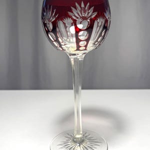 Puede incluir: Una copa de vino transparente con un cuenco rojo rubí y un tallo largo y delgado. El cuenco presenta intrincados diseños florales y geométricos blancos grabados. La base es un patrón claro de estrella. La copa está sobre un fondo blanco.