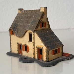 May include: A miniature model of a cottage with cream-colored walls, a textured dark gray thatched roof, and orange shutters. The structure features multiple windows, two doors, and stone-like detailing around the edges, all set on a black base.