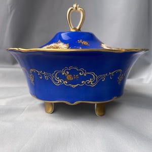 Może przedstawiać: Kobaltowa miska porcelanowa z pasującą pokrywką, ozdobiona złotymi motywami kwiatowymi i wykończeniami. Misa wspiera się na trzech małych, złoconych nóżkach i posiada centralny, złoty uchwyt umieszczony na pokrywce.