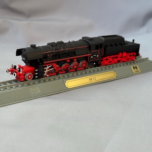 May include: A black model steam locomotive with red wheels and undercarriage, mounted on a gray plastic display track base. A gold-colored plaque on the front of the base features the text BR 52, and a small square N scale logo is visible.