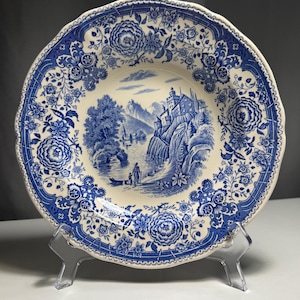 Könnte beinhalten: Ein blau-weißer Zierteller mit floralem Rand und einer zentralen Landschaftsszene. Der Teller zeigt ein detailliertes Design eines Schlosses, Bäumen und Figuren in der Nähe eines Gewässers. Der Teller wird auf einem transparenten Ständer ausgestellt.