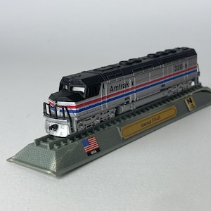 Könnte beinhalten: Eine silberne Amtrak FP-45 Modell-Lokomotive mit schwarzem Dach und Front, verziert mit roten und blauen horizontalen Streifen. Der Zug ist auf einem grauen Sockel montiert, der eine goldene Plakette mit der Aufschrift Amtrak FP-45 trägt.