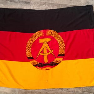 May include: A rectangular flag featuring horizontal stripes of black, red, and gold. Centered on the red stripe is a circular emblem depicting a yellow hammer and compass surrounded by a golden wreath of wheat stalks on a red background.