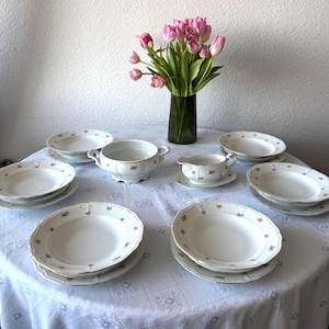 Könnte beinhalten: Ein Vintage-Tafelservice aus weißem Porzellan mit floralem Design, einschließlich Teller, einer Terrine und einer Sauciere. Das Set ist auf einer weißen Tischdecke angeordnet, mit einer Vase mit rosa Tulpen im Hintergrund.