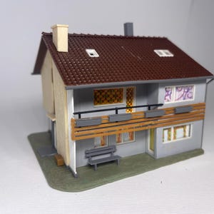 Könnte beinhalten: Ein Modellhaus mit zwei Stockwerken, einem braunen Ziegeldach, einer hellgrauen Fassade und einer beigen Seitenwand. Das Gebäude verfügt über einen Balkon mit Holzgeländer, grauen Blumenkästen, einer kleinen Bank und mehreren Fenstern.