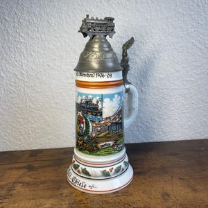 Può includere: Un boccale da birra in ceramica con coperchio in metallo sormontato da una locomotiva in miniatura. Il corpo presenta un'illustrazione colorata di un treno, il testo "München 1906-09", "meine", "Dienstzeit" e una base decorata con foglie di quercia.
