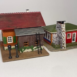 Könnte beinhalten: Zwei Miniatur-Modellgebäude. Das erste ist ein zweistöckiges Bauwerk mit rotem Ziegeldach, orangefarbenen Ziegelwänden, grünen Fensterläden und einem grau gedeckten Anbau mit schwarzer Leiter. Das zweite ist ein rotes Gebäude mit grünem Dach.