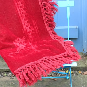 May include: A rich red velvet throw blanket featuring a floral design and fringed edges. The blanket is draped over a light blue metal chair. The contrast between the red and blue creates a visually appealing scene. A home decor item.