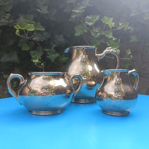 May include: A three-piece silver-toned metal tea set consisting of a large pitcher with a handle, a wide sugar bowl with two side handles, and a smaller creamer pitcher with a single handle. Each piece features a highly reflective, polished surface.