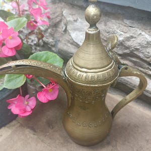 May include: A brass coffee pot with a long, curved spout and a tapered lid topped with a spherical finial. The body features intricate engraved patterns around the neck and base, a curved handle, and a hinged lid mechanism on the upper section.