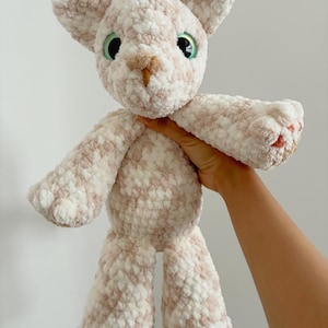 May include: A handmade crochet cat plushie in cream and light brown. The cat has green eyes, a tan nose, and outstretched arms. The plush toy is soft and fluffy, with a textured appearance.