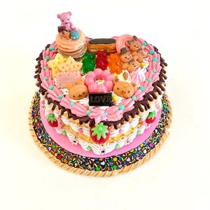May include: A heart-shaped cake decorated with a variety of sweets, including gummy bears, cookies, doughnuts, and chocolate. The cake is topped with a pink teddy bear and the word "LOVE" in chocolate.
