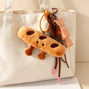 May include: A cream-coloured canvas tote bag featuring a plush, fuzzy tan cookie-shaped keychain with dark brown spots, a smiling face, and two small legs. The keychain includes a brown cord, a yellow ring, and a glossy, light brown candy charm.