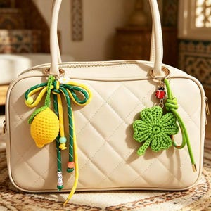 May include: A cream-colored quilted handbag with two top handles. The bag is decorated with two charms: one featuring a yellow crocheted lemon with a green bow and beads, and the other a green crocheted four-leaf clover with a small ladybug charm.