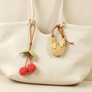 May include: A smooth off-white tote bag featuring two hanging crochet charms. One charm is a pair of red cherries with green leaves on a brown cord. The second charm is a miniature beige crochet bag with floral details on a brown cord.