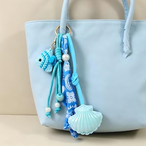 May include: A light blue handbag featuring a decorative keychain accessory. The keychain includes a small crocheted blue fish, a blue and white patterned fabric ribbon, several white beads, and a light blue plastic seashell charm.