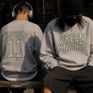 Pode incluir: Duas pessoas usando moletons cinza com texto e números sentadas em um banco em uma quadra de basquete. Um moletom diz "WESTBERG 11" e o outro "GREEN KNIGHTS". Uma bola de basquete e tênis brancos em primeiro plano.