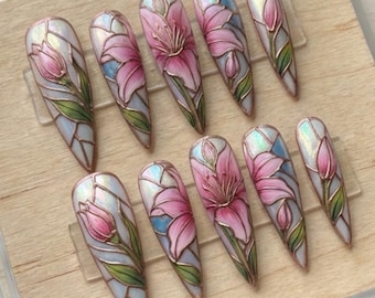 May include: A set of ten long, almond-shaped press-on nails. Each nail features a pink lily design with gold outlines, set against a stained-glass effect background with blue and white hues. The nails are displayed on a wooden base within a clear acrylic box.