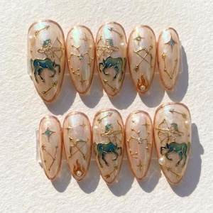 Puede incluir: Juego de diez uñas postizas en forma de almendra con acabado nacarado. Cada uña presenta un diseño dorado de Sagitario con un centauro turquesa, arco y flecha, y detalles de constelación. Algunas uñas tienen pequeñas perlas y detalles de llama.