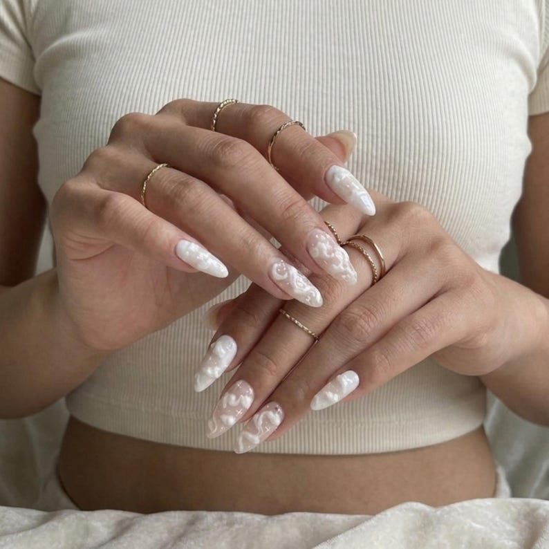 May include: Long, white, stiletto-shaped artificial nails with a 3D floral design. The nails are adorned with delicate, raised white floral patterns. Several gold rings are worn on the fingers. The nails are a fashionable accessory.