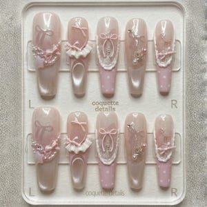 May include: A set of ten long, light pink press-on nails with various designs. Some nails feature bows, pearls, and lace details. The nails are arranged in a clear plastic tray, with the text "coquette details" visible.